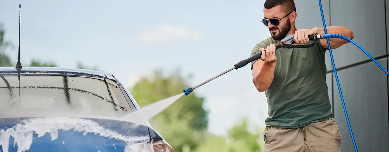 A man uses a pressure washer to clean his car, spraying water to remove dirt and grime from the vehicle's surface.