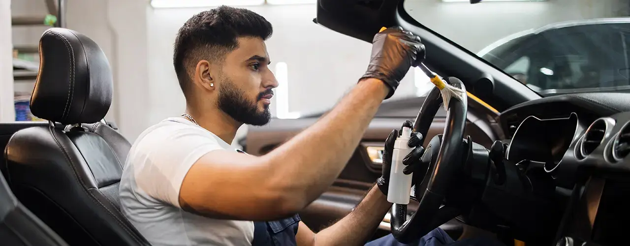 A man seated in the driver's position, cleaning the inside of the car.