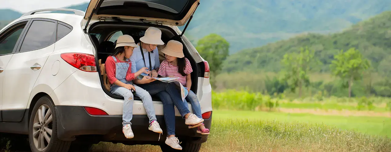 Young family sitting in the trunk of their car, surrounded by greenery and enjoying a sunny spring day.