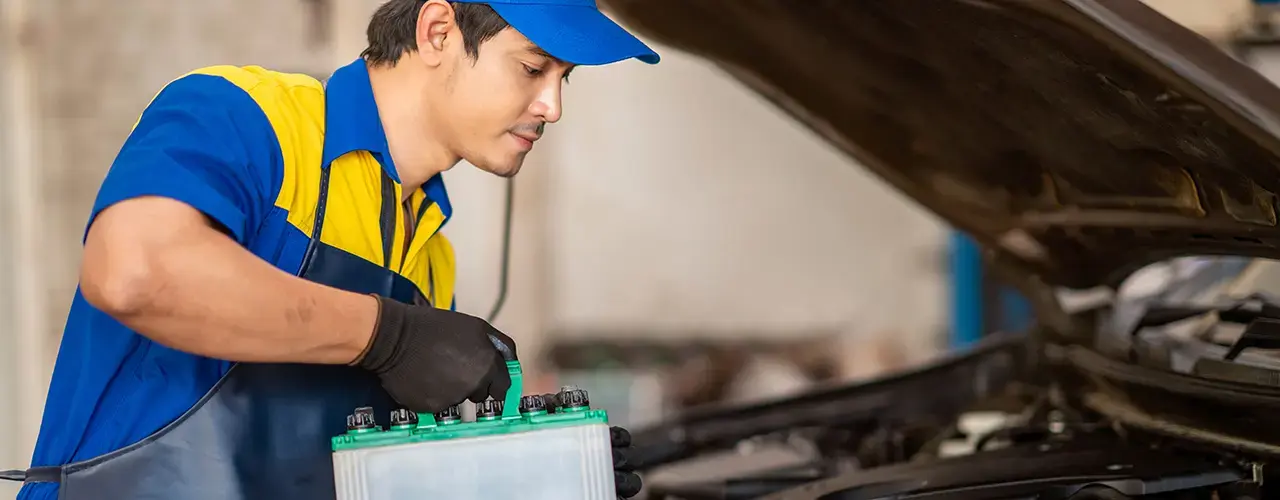 A mechanic in a blue shirt and hat is replacing a car battery.