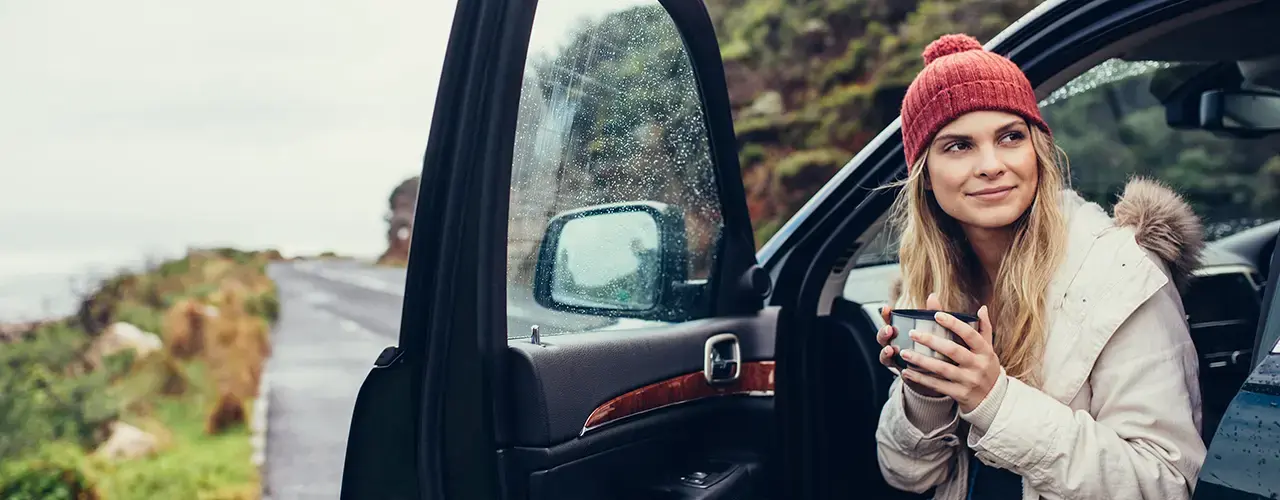 A woman in a car, bundled in winter attire, enjoys a cup of coffee.